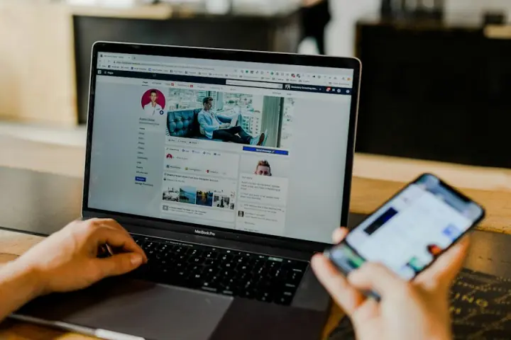 Eine Person tippt auf einem Laptop mit Facebook-Profil und hält ein Smartphone.